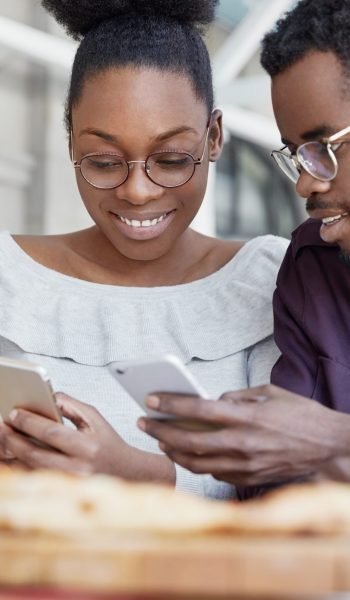 Happy African American couple of enterpreneurs develop new business strategy on portable laptop computer, use mobile phones for surfing information in internet, sit together in spacious office room